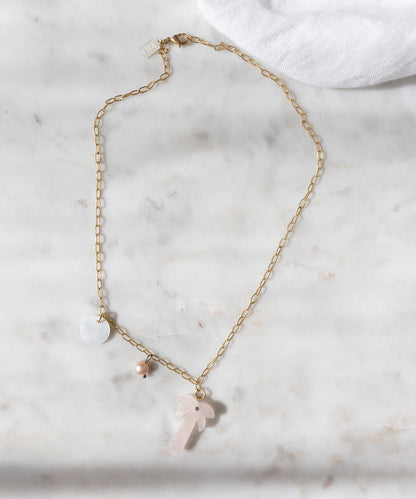 The WALD Berlin Mexicana Solo Amore Necklace is a handmade gold-plated piece with three pendants—a white disc, small bead, and light pink hand-carved stone palm—shown elegantly on marble.
