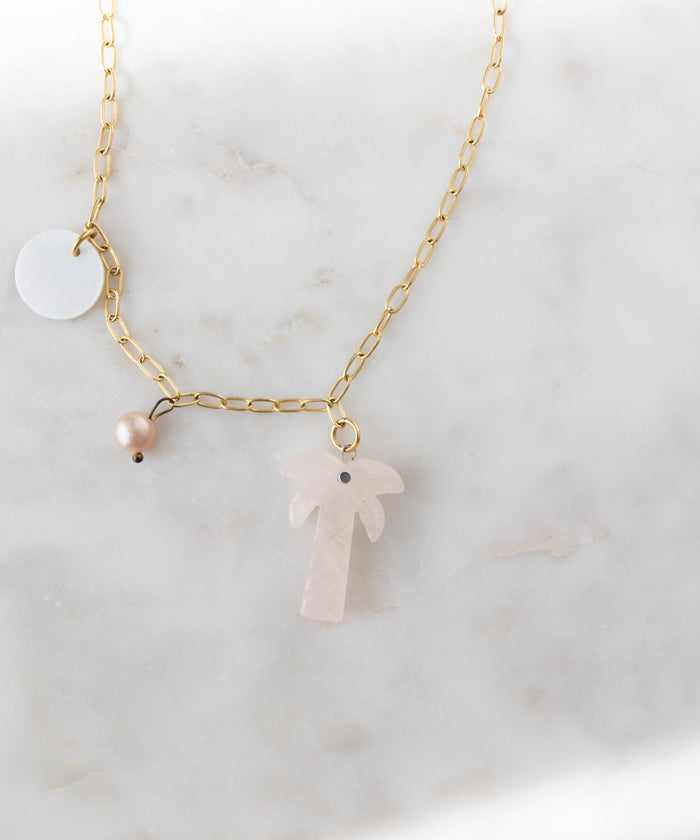 The WALD Berlin Mexicana Solo Amore Necklace is a handmade gold-plated piece with a hand-carved stone palm, a white round charm, and a pink pearl charm, showcased on white marble.