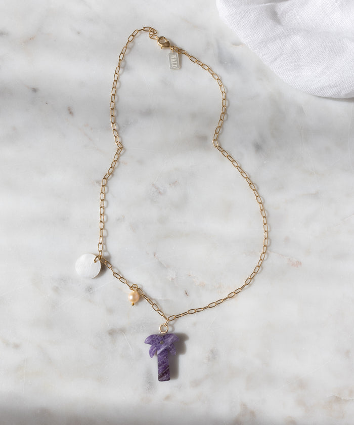 The WALD Berlin Mexicana Solo Amore Necklace is a handmade gold-plated piece with a purple stone pendant, a round white charm, and a small pearl, elegantly displayed on white marble.