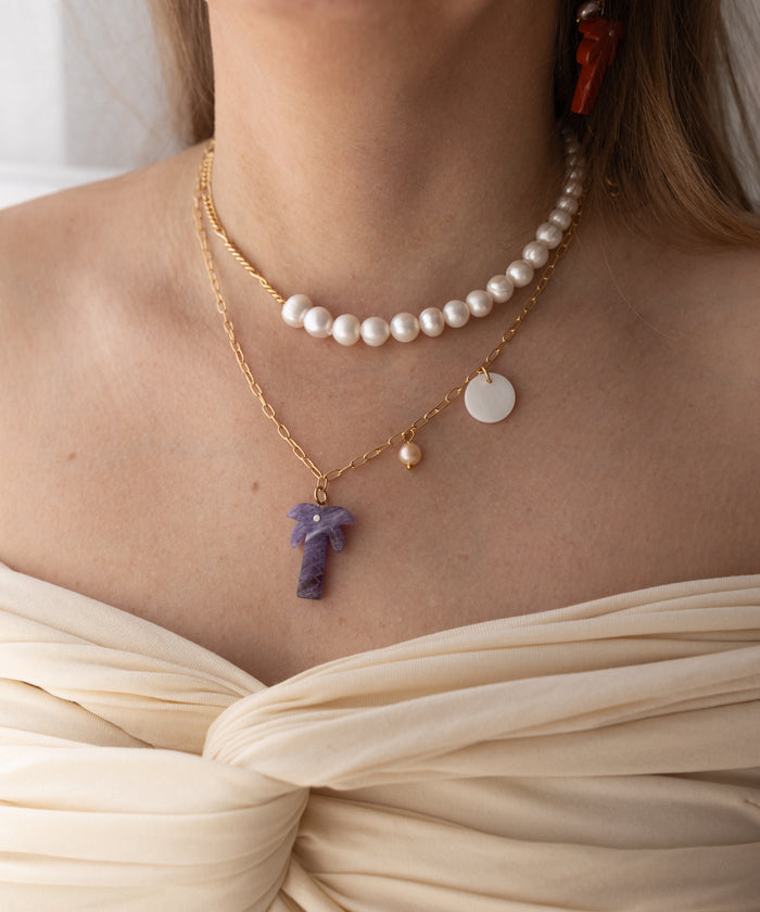 A woman in an off-shoulder cream top wears layered necklaces: a pearl choker, WALD Berlin's Mexicana Solo Amore Necklace with a handmade gold-plated chain, round pendant and pearl, and a gold chain with a purple stone pendant.