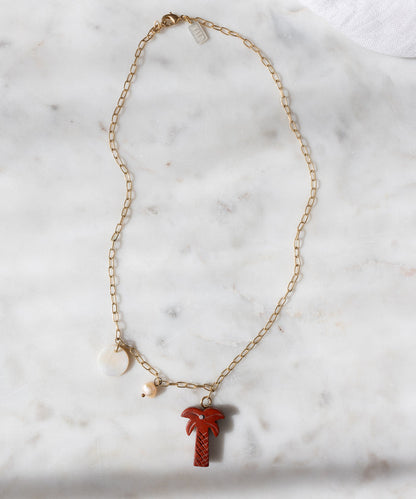 The WALD Berlin Mexicana Solo Amore Necklace is handmade, gold-plated, and showcases a hand-carved stone palm pendant, a small white pearl, and a round white charm on a white marble surface.