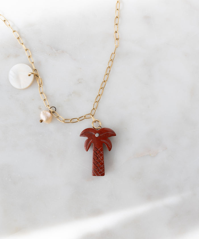 The WALD Berlin Mexicana Solo Amore Necklace is a handmade gold-plated piece with a hand-carved stone palm pendant, a round white charm, and a pearl charm, elegantly showcased on white marble.