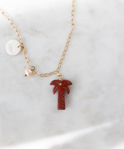 The WALD Berlin Mexicana Solo Amore Necklace is a handmade gold-plated piece with a hand-carved stone palm pendant, a round white charm, and a pearl charm, elegantly showcased on white marble.