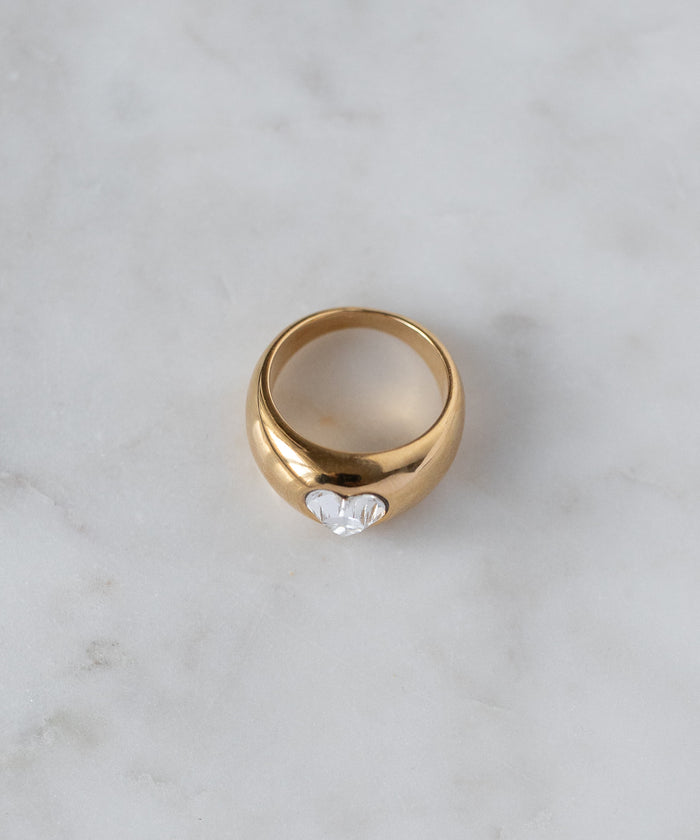 The WALD Berlin Mom Swarovski Heart Ring, gold plated with a single clear Swarovski stone at the center, is displayed on a light marble surface.