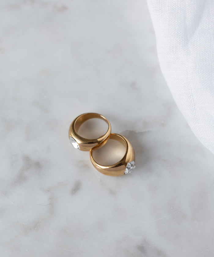 The WALD Berlin Mom Swarovski Heart Ring, a gold plated piece with a clear gemstone, is displayed on light marble beside soft white fabric.