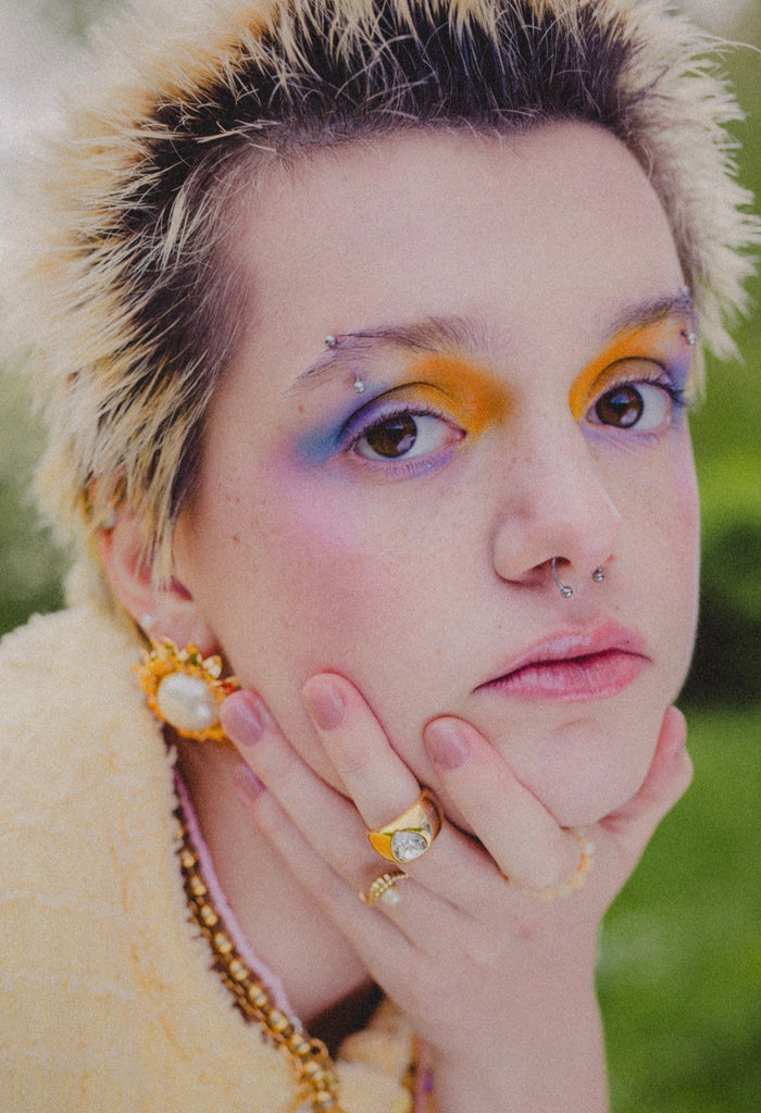 Person with short, spiky blond hair and colorful eye makeup rests their chin on their hand, wearing WALD Berlin's Mom Swarovski Heart Ring and gold jewelry, looking directly at the camera.