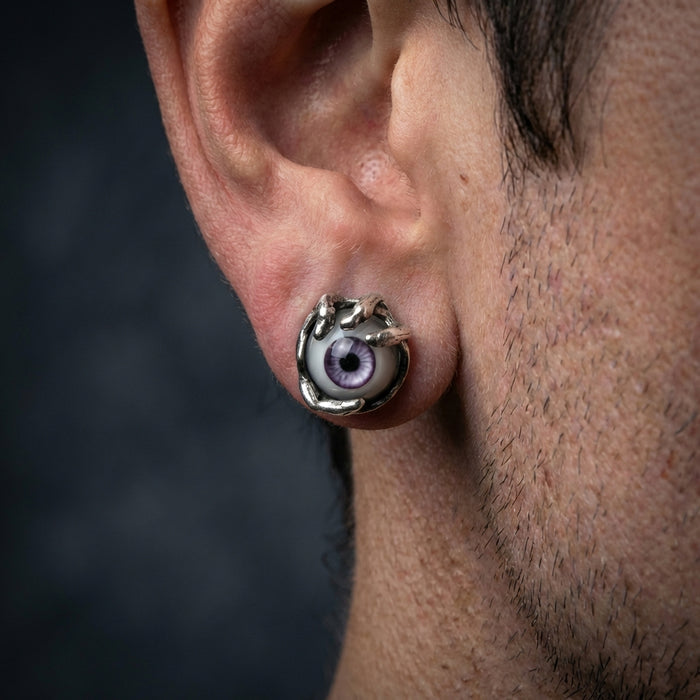 Silver monster eye earring with purple iris and claw setting worn on a man's ear, featuring gothic horror-inspired design with realistic eyeball detail