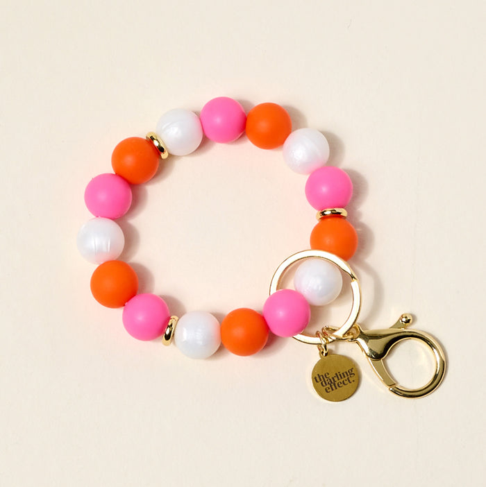 The Darling Effect's On the Brightside Hands-Free Keychain Wristlet features pink, orange, and white silicone beads, a gold clasp, keyring, and a "the darling effect" charm—keeping your keys stylish and hands-free.