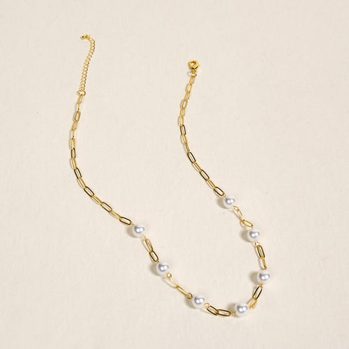 The Darling Effect Pearl Paperclip Necklace Chain, featuring elongated links, spaced white pearls, and delicate charms, is displayed on a light beige surface.
