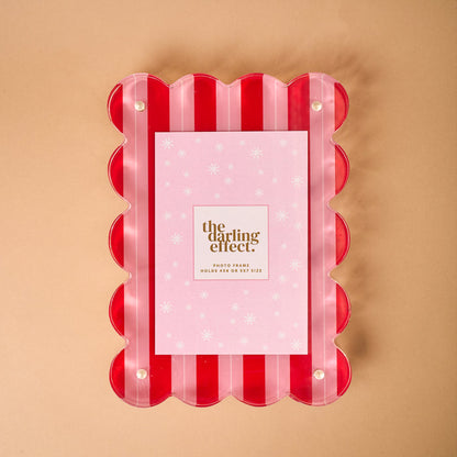 The Peppermint Stripes Rectangle Scalloped Acrylic Frame by The Darling Effect features pink and red stripes with scalloped edges. It holds 5x7" (13x18cm) photos and is perfect for modern home décor. Displayed on a beige surface with a branded placeholder card.