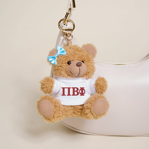 The Darling Effect's Pi Beta Phi Teddy Bear Bag Charm, featuring a blue bow and a white shirt with red Greek letters, hangs on a stylish white handbag.
