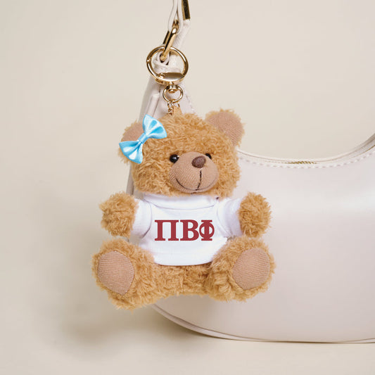The Darling Effect's Pi Beta Phi Teddy Bear Bag Charm, featuring a blue bow and a white shirt with red Greek letters, hangs on a stylish white handbag.