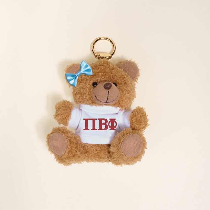 The Darling Effect’s Pi Beta Phi Teddy Bear Bag Charm is a small brown bear keychain with a blue bow, dressed in a white shirt with red ΠΒΦ Greek letters—perfect for Bid Day—shown on a light beige background.