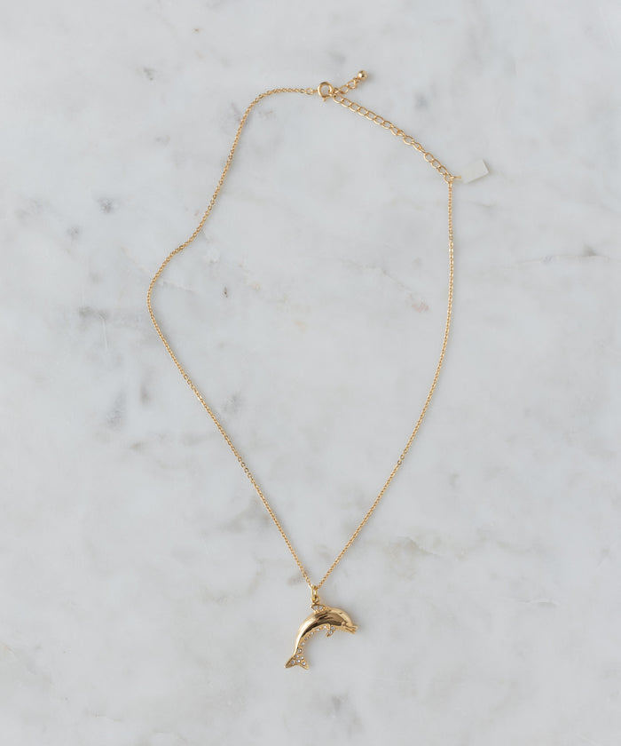 The WALD Berlin Pop Dolphin Necklace features a dolphin-shaped pendant in gold-plated brass, adorned with sparkling Swarovski stones, and is shown on a light marble surface.