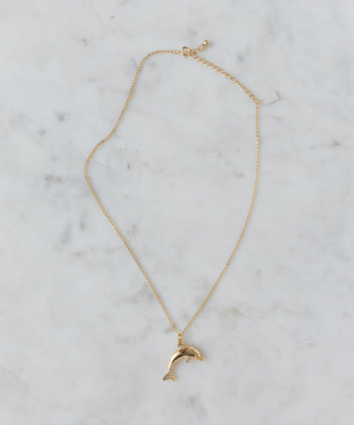 The WALD Berlin Pop Dolphin Necklace features a dolphin-shaped pendant in gold-plated brass, adorned with sparkling Swarovski stones, and is shown on a light marble surface.