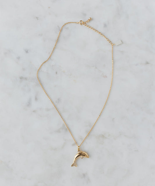 The WALD Berlin Pop Dolphin Necklace features a dolphin-shaped pendant in gold-plated brass, adorned with sparkling Swarovski stones, and is shown on a light marble surface.