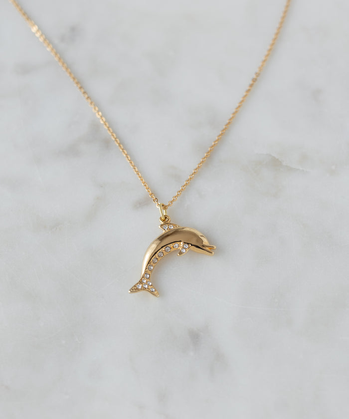 The WALD Berlin Pop Dolphin Necklace, a goldplated brass piece with a dolphin-shaped pendant embellished with sparkling Swarovski stones, is elegantly displayed on a light marble surface.
