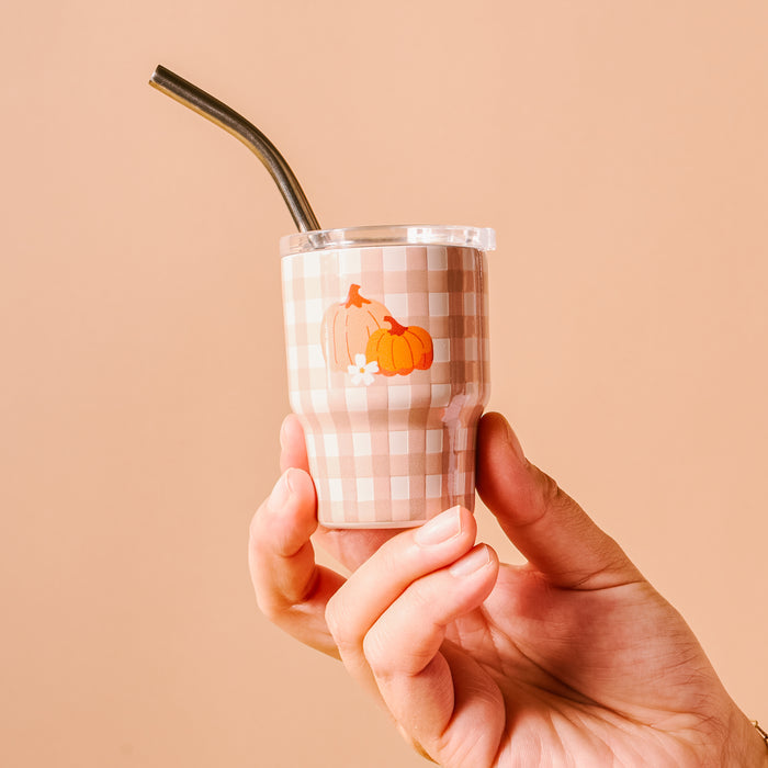 A hand holds The Darling Effect Pumpkin Gingham 2 oz Tiny Tumbler, featuring a beige and white checkered pattern, two pumpkin decals, and a metal straw—perfect for autumn party favors.