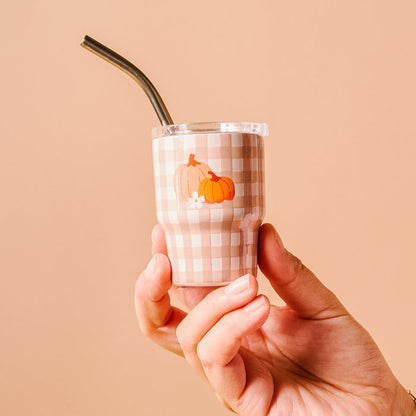 A hand holds The Darling Effect Pumpkin Gingham 2 oz Tiny Tumbler, featuring a beige and white checkered pattern, two pumpkin decals, and a metal straw—perfect for autumn party favors.