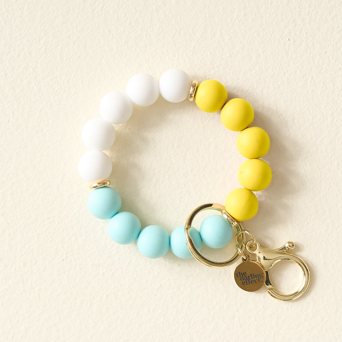 The Darling Effect's Ray of Sunshine Hands-Free Keychain Wristlet features white, yellow, and light blue silicone beads, gold key rings, and a charm engraved with The Giving Effect—perfect for holding keys stylishly on the go.