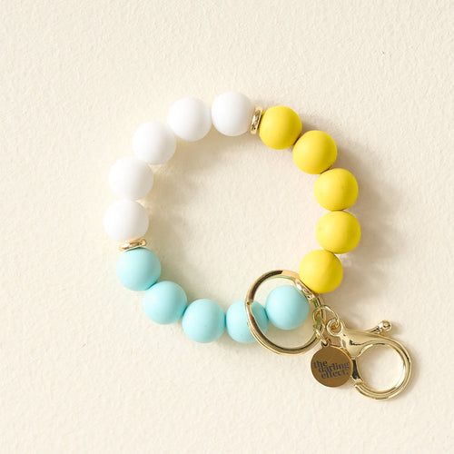 The Darling Effect's Ray of Sunshine Hands-Free Keychain Wristlet features white, yellow, and light blue silicone beads, gold key rings, and a charm engraved with The Giving Effect—perfect for holding keys stylishly on the go.