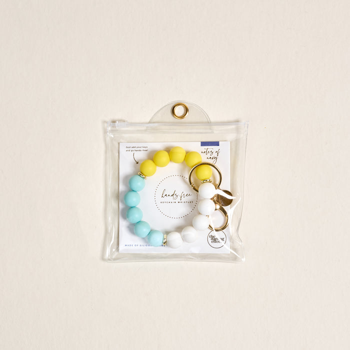 The Darling Effect's Ray of Sunshine Hands-Free Keychain Wristlet, featuring yellow, blue, and white beads, is displayed in a clear plastic pouch with a gold grommet against a light beige background.