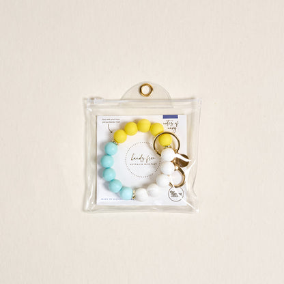 The Darling Effect's Ray of Sunshine Hands-Free Keychain Wristlet, featuring yellow, blue, and white beads, is displayed in a clear plastic pouch with a gold grommet against a light beige background.