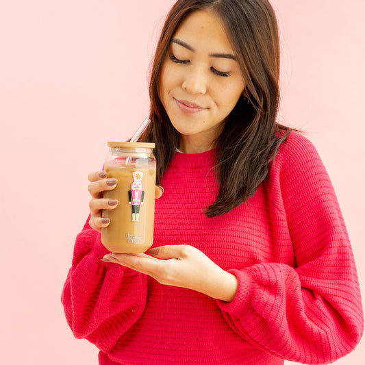 A woman in a bright pink sweater holds The Darling Effect’s Round Glass Cup - Holiday Check, filled with iced coffee and topped with a bamboo lid, featuring a festive nutcracker sticker and straw. She smiles while looking down at the drink, set against a pink background.