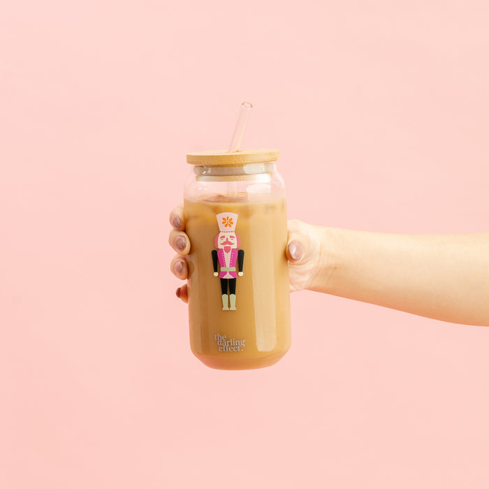 A hand holds The Darling Effect’s Round Glass Cup - Holiday Check, a glass cup of iced coffee with a bamboo lid and straw. The cup features a festive check pattern printed on it, set against a pink background.