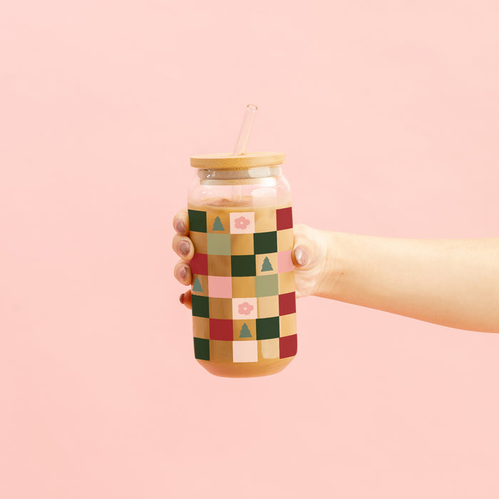 A hand holds The Darling Effect’s Round Glass Cup - Holiday Check, which features a bamboo lid and straw and is decorated with a pink, green, and yellow patchwork pattern with trees and flowers, set against a light pink background.