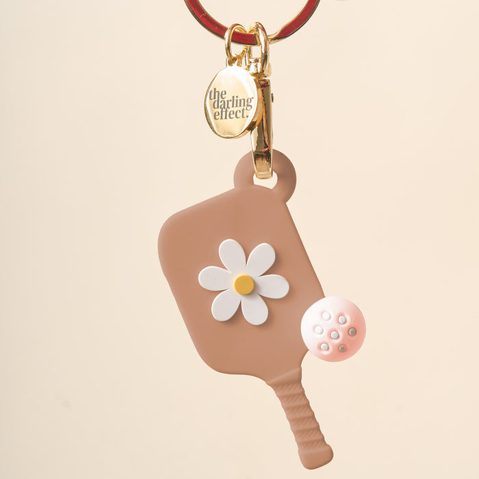 The Darling Effect Sandstone Pickleball Paddle Keychain features a brown paddle with a white and yellow flower design, gold keyring, pink pickleball charm, and makes the perfect unique gift for pickleball fans.