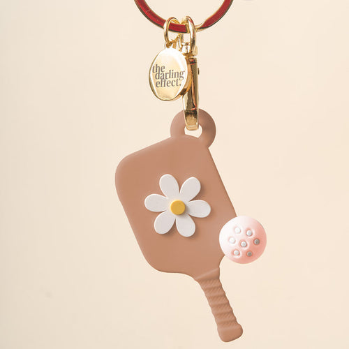 The Darling Effect Sandstone Pickleball Paddle Keychain features a brown paddle with a white and yellow flower design, gold keyring, pink pickleball charm, and makes the perfect unique gift for pickleball fans.
