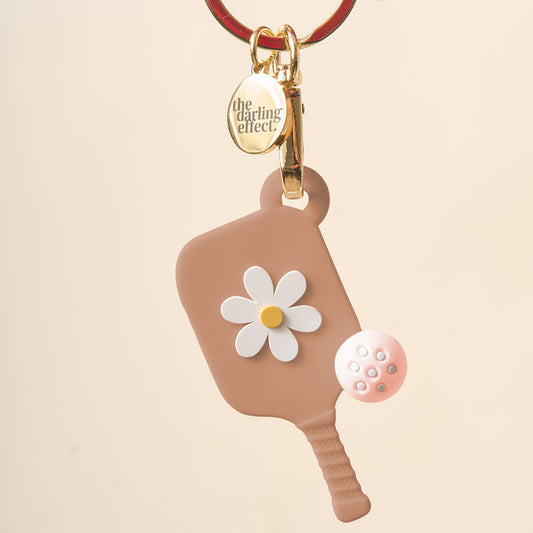 The Darling Effect Sandstone Pickleball Paddle Keychain features a brown paddle with a white and yellow flower design, gold keyring, pink pickleball charm, and makes the perfect unique gift for pickleball fans.