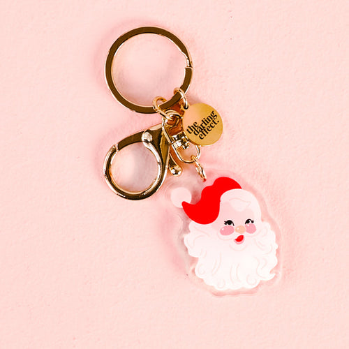 The Darling Effect’s Santa Acrylic Keychain features a cute, cartoon-style Santa face charm with a red hat on a gold clip and round tag. Made from durable acrylic, it’s shown on a light pink background.