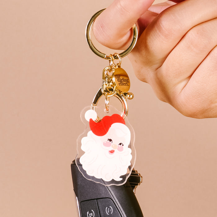 A hand holds a car key with The Darling Effect's Santa Acrylic Keychain—a durable, festive accessory featuring a smiling Santa Claus in a red hat on a gold keyring.