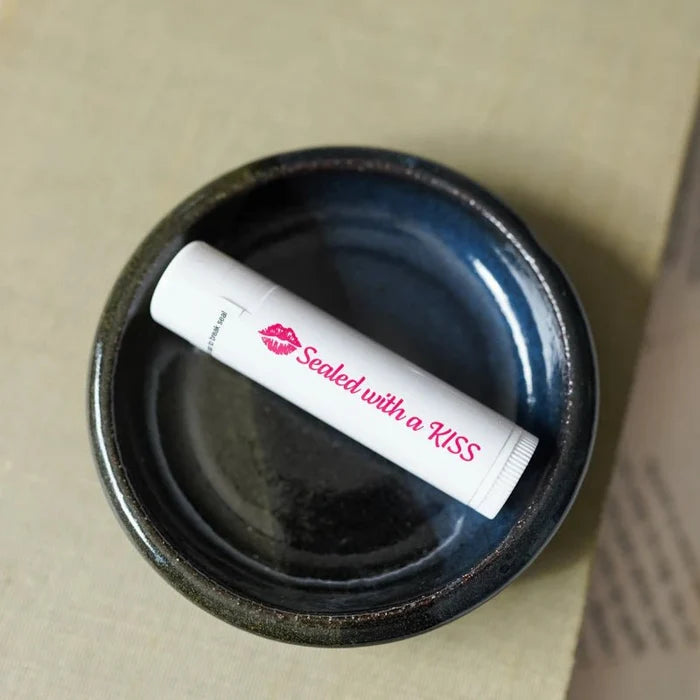 Sealed with a Kiss lip balm from LitJoy Winter Romantasy TBR Box — white lip balm tube with pink kiss print and cursive text resting in a dark blue ceramic dish
