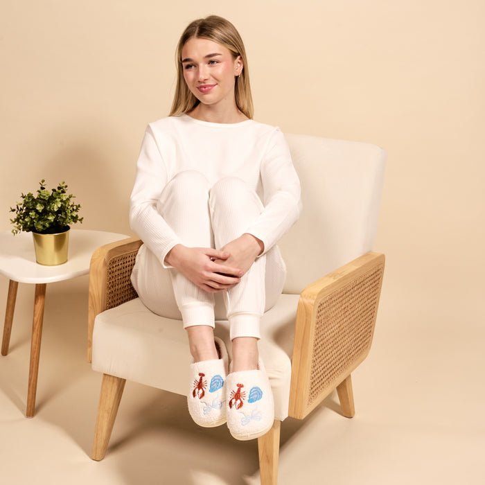 A woman in white loungewear, smiling and cross-legged in a beige armchair, wears The Darling Effect's Seaside Haven Fuzzy Slippers with blue and red embroidery. Beside her is a small plant on a white table against a beige background.