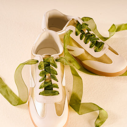 Two white sneakers with gold accents are styled with The Darling Effect’s Sheer Green Step It Up Shoelaces, set against a light beige background.