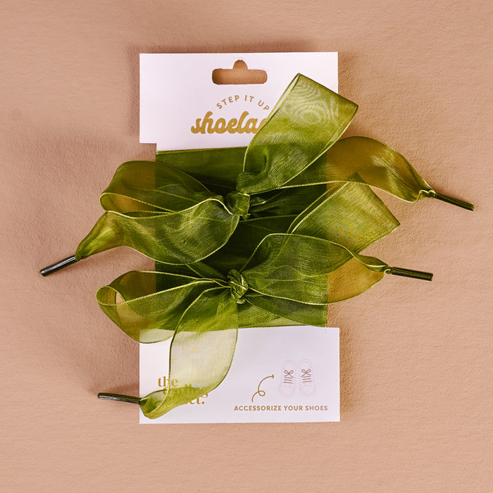 A pair of Sheer Green Step It Up Shoelaces by The Darling Effect are tied in bows and displayed on a white backing card with shoe illustrations, set against a beige background.