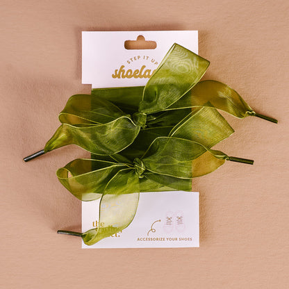 A pair of Sheer Green Step It Up Shoelaces by The Darling Effect are tied in bows and displayed on a white backing card with shoe illustrations, set against a beige background.