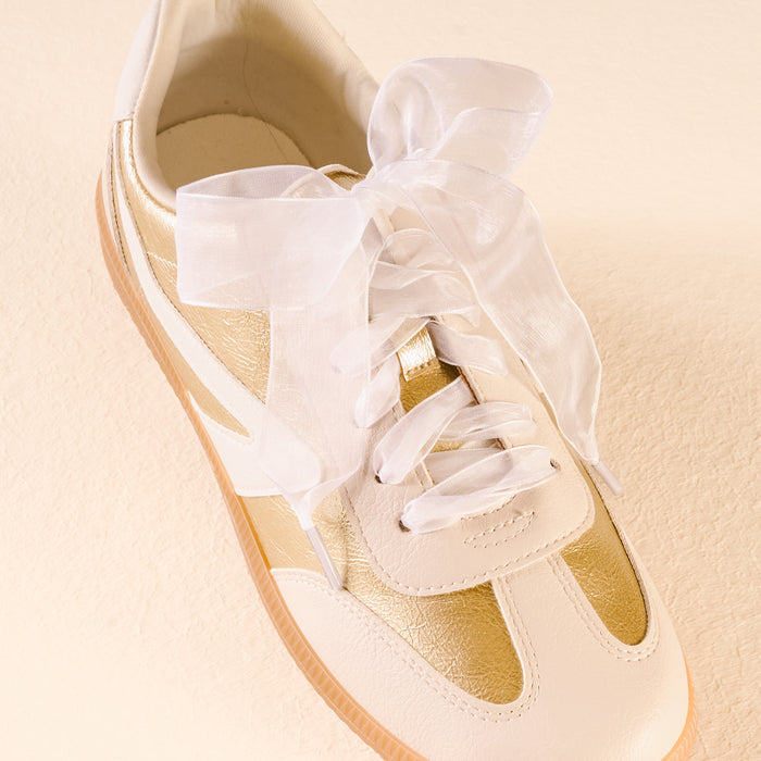 The Darling Effect’s Sheer White Step It Up Shoelaces are large, sheer white organza laces tied in a bow on a white and gold sneaker with a gum sole, shown against a light beige background.