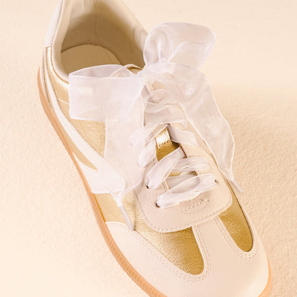 The Darling Effect’s Sheer White Step It Up Shoelaces are large, sheer white organza laces tied in a bow on a white and gold sneaker with a gum sole, shown against a light beige background.