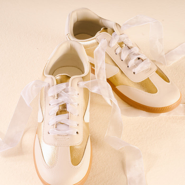 A pair of gold and cream sneakers are laced with Sheer White Step It Up Shoelaces by The Darling Effect, neatly tied against a pale yellow background.