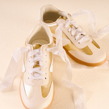 A pair of gold and cream sneakers are laced with Sheer White Step It Up Shoelaces by The Darling Effect, neatly tied against a pale yellow background.