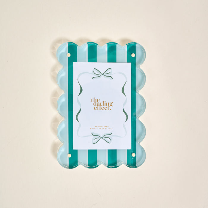 The Sicily Stripes Teal Rectangle Scalloped Acrylic Picture Frame by The Darling Effect features alternating teal and clear vertical stripes, holding a gold-lettered card, displayed on a light beige background.