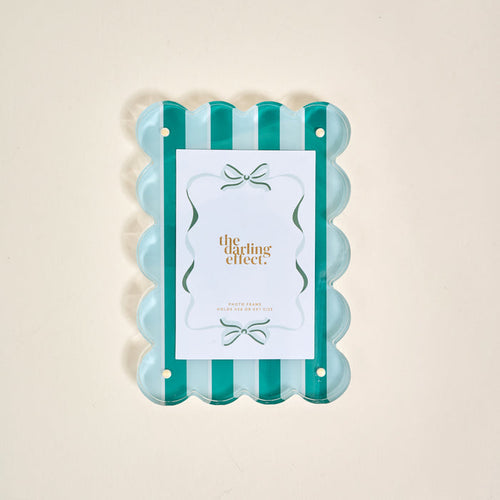 The Sicily Stripes Teal Rectangle Scalloped Acrylic Picture Frame by The Darling Effect features alternating teal and clear vertical stripes, holding a gold-lettered card, displayed on a light beige background.