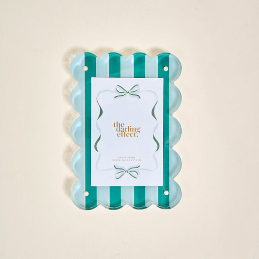 The Sicily Stripes Teal Rectangle Scalloped Acrylic Picture Frame by The Darling Effect features alternating teal and clear vertical stripes, holding a gold-lettered card, displayed on a light beige background.