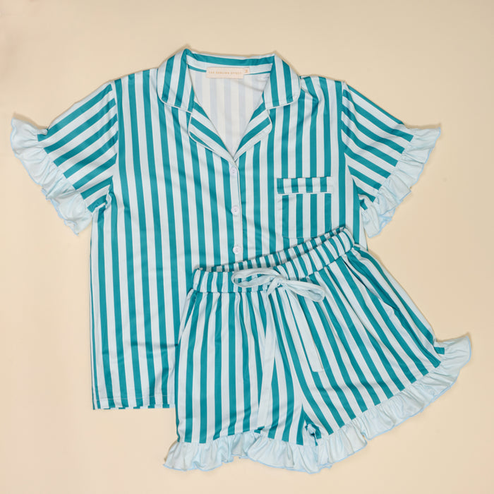 The Darling Effect’s Sicily Stripes Teal Slumber Party Pajama Shorts Set features teal and white stripes on ultra-soft fabric, with a short-sleeved ruffled button-up top and high-waisted shorts with a tie belt and ruffled hems, laid flat on beige.