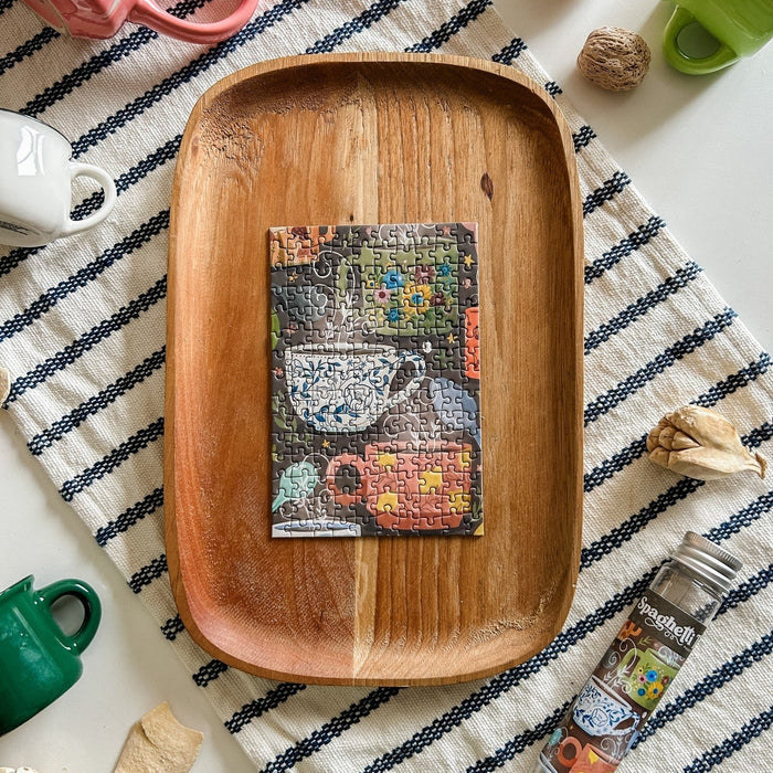 The Sip & Savor | 150 Piece Jigsaw Puzzle by Spaghetti Tree features a teacup image completed on a wooden tray, set amid cups and kitchen items—a delightful, portable scene for puzzle fans.
