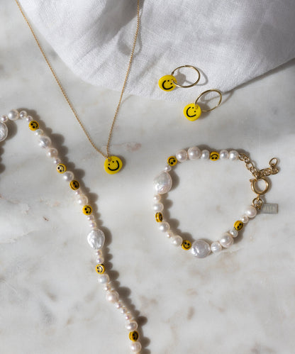 Eine Perlenkette und ein Armband, verziert mit gelben Smiley-Perlen, eine goldene Kette sowie die „Smilie Yellow“-Ohrringe von WALD Berlin – vergoldete Creolen mit passenden, in Deutschland gefertigten Smiley-Anhängern – präsentiert auf einer weißen Marmorfläche.